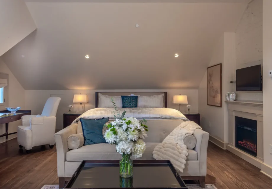 Elegant bedroom with sloped ceiling, plush bedding, fireplace, armchair, and TV.
