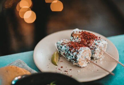 two elote corn cobs on sticks with lime wedge on cream colored plate on blue surface