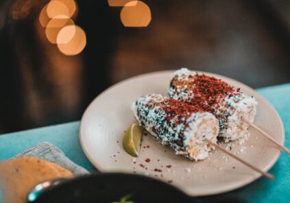 two elote corn cobs on sticks with lime wedge on cream colored plate on blue surface