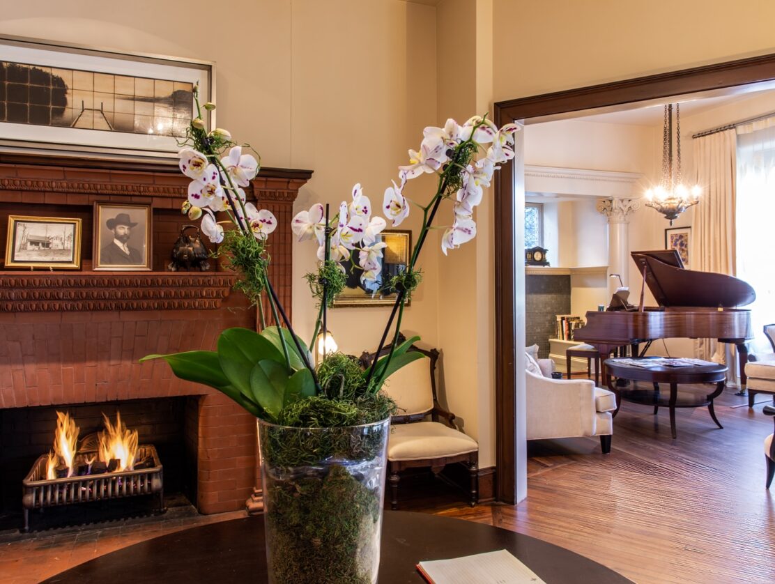 White orchids in glass vase on table in front of fire place and living room with white couches and brown piano