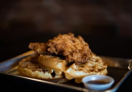 chicken and waffles on dark plate with dark background and syrup cup in foreground